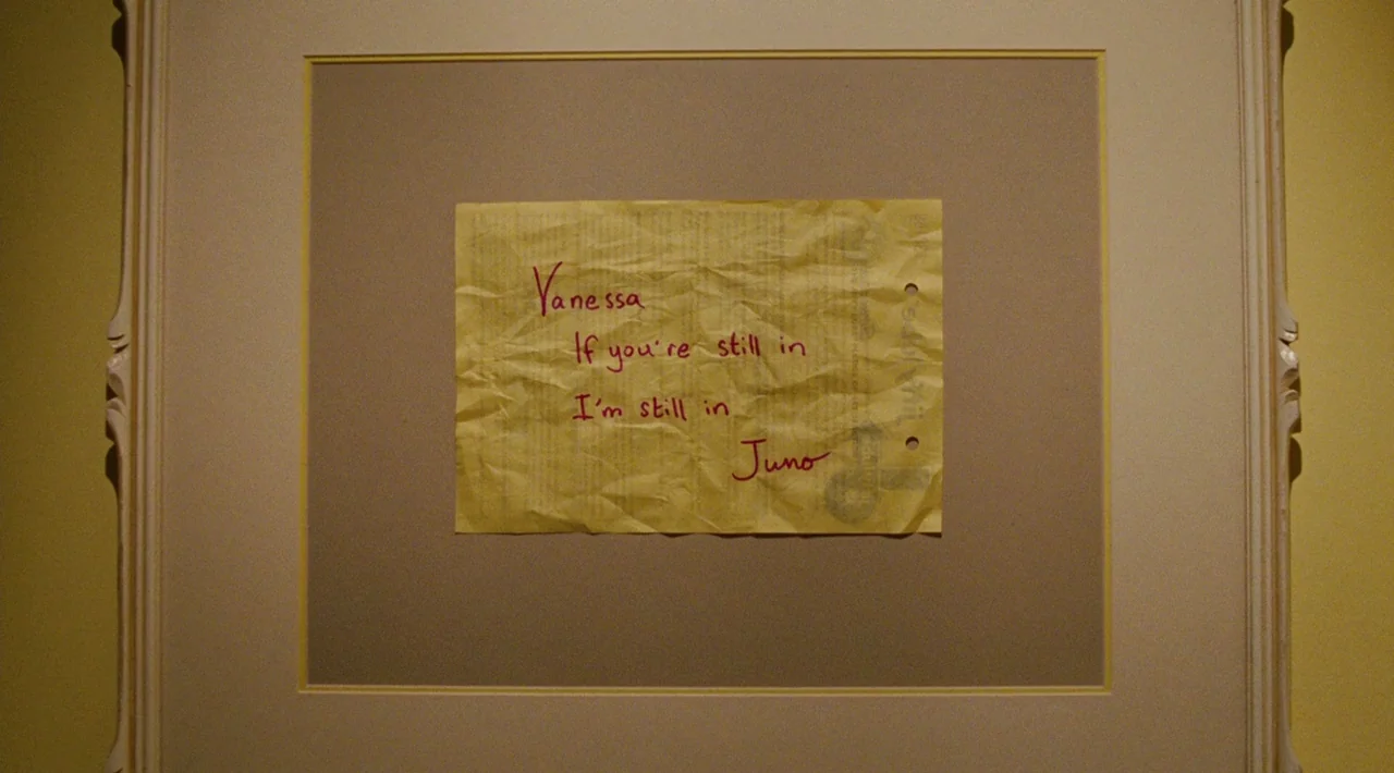 Juno (2007) movie still: closeup — A framed piece of wrinkled yellow paper hangs on a plain cream-colored wall. The paper contains a handwritten…