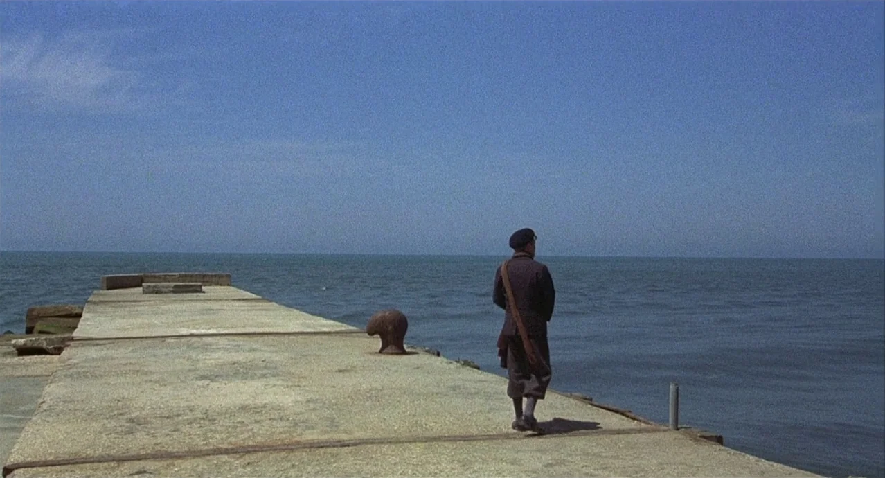 Amarcord (1973) movie still: wide — A concrete pier extends into calm water under a clear blue sky. A man in dark clothing walks away from the…