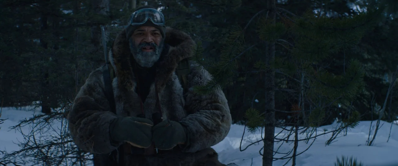 Hold The Dark (2018) movie still: medium — A man stands in a dense, snow-covered forest during a cold, overcast day. The subject wears heavy winter…