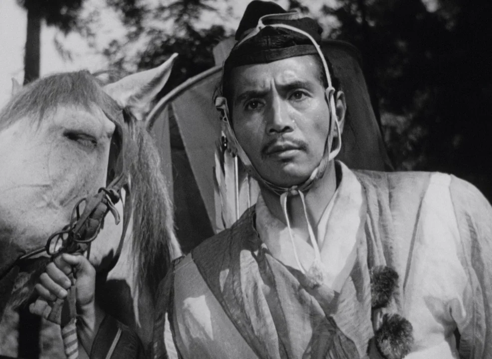 Rashomon (1950) movie still: medium — A man in traditional robes stands next to a horse outdoors. The background displays blurred foliage and a tall…