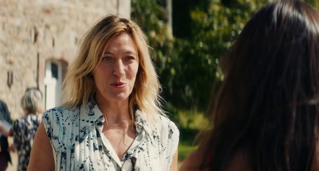 Anaïs in Love (2021) movie still: medium — An outdoor area features a stone wall and blurred greenery in the background. A woman in a patterned…