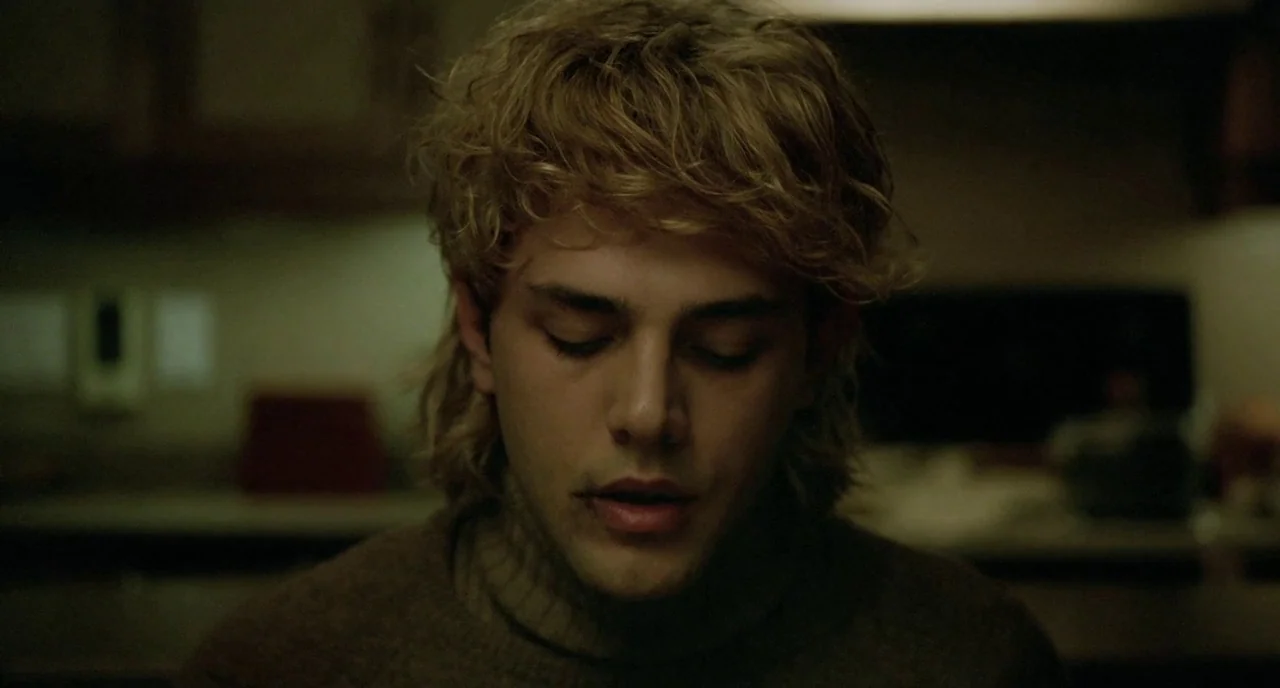 Tom at the Farm (2013) movie still: closeup — A dimly lit room with out-of-focus household objects in the background. A young man with blonde curly…