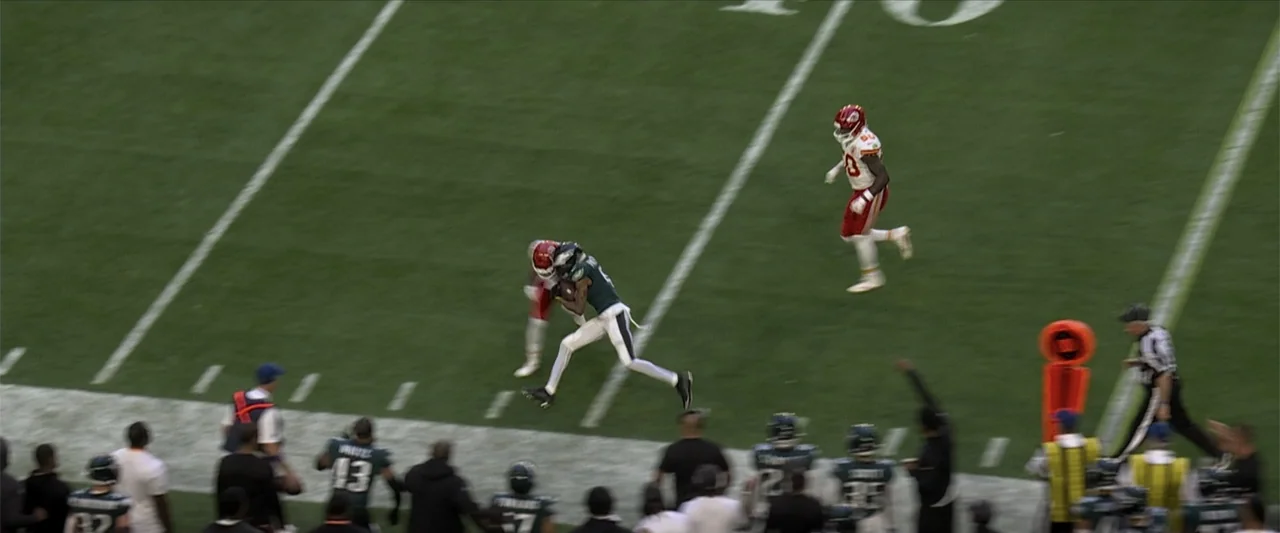 Kelce (2023) movie still: wide — A professional football field at night with marked yard lines and painted grass. Two players in contrasting team…