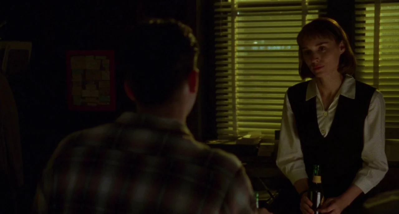 Carol (2015) movie still: medium — A dimly lit bar interior features windows covered with yellow horizontal blinds at night. A woman stands behind…