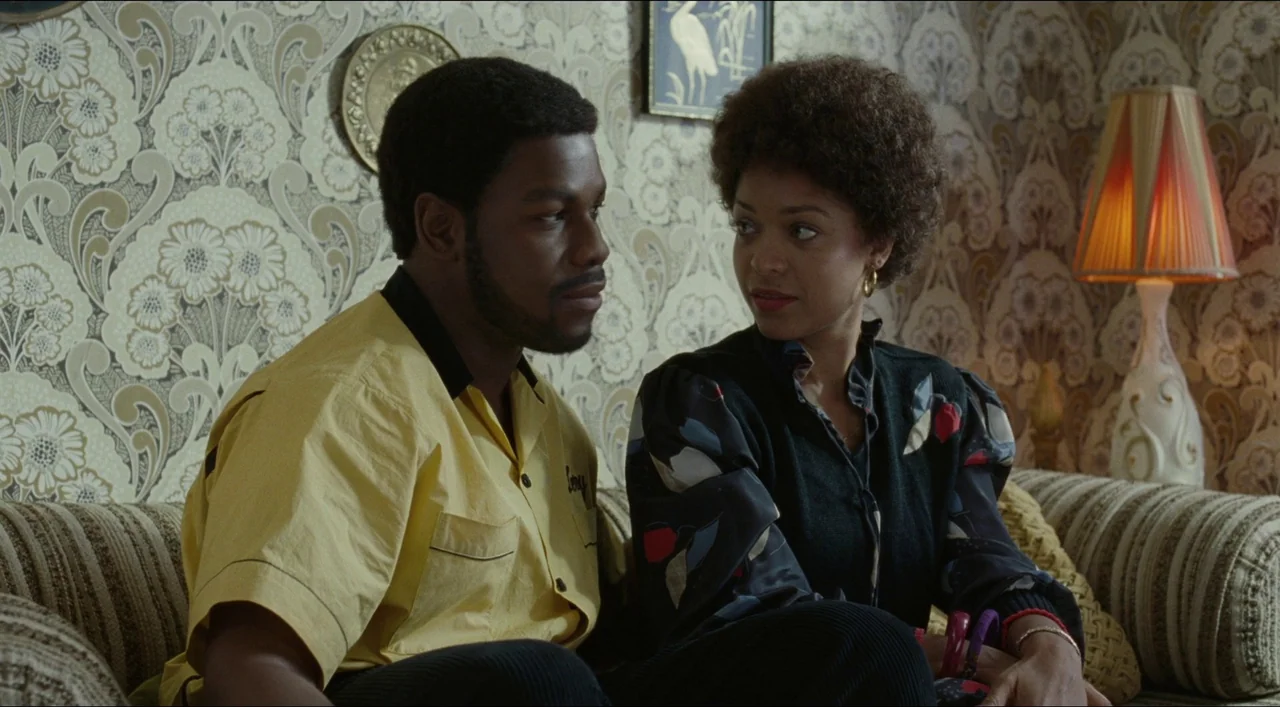 Red, White & Blue (2020) movie still: medium — A 1970s living room features patterned tan wallpaper and a lit table lamp. A man and a woman sit side…