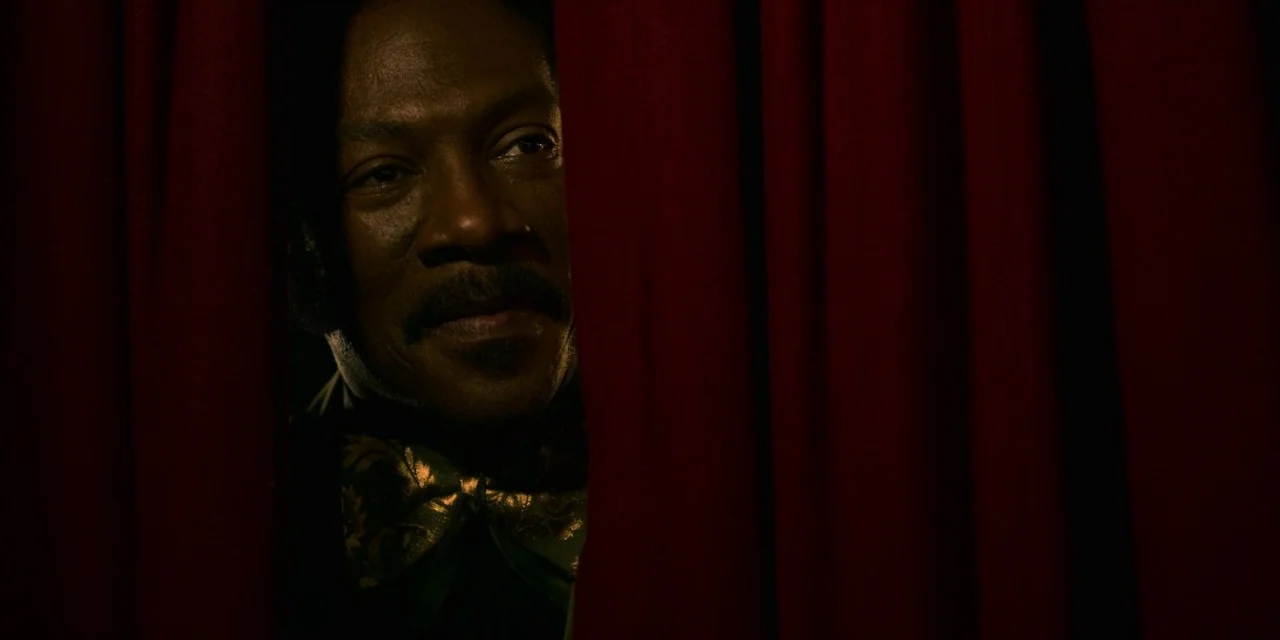 Dolemite Is My Name movie still: closeup — A man peers through a narrow opening between two heavy velvet red curtains. He wears a dark shirt and a…