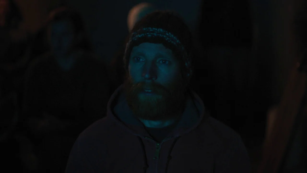 Night Moves (2013) movie still: closeup — A dark, dimly lit environment with indistinct figures visible in the background. A man wearing a beanie…