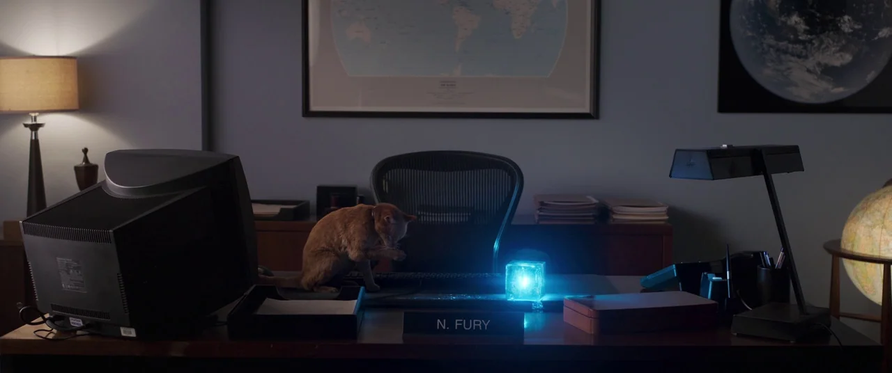 Captain Marvel (2019) movie still: wide — A dark wooden desk sits in a quiet office with a world map on the wall. A ginger tabby cat sits on the…