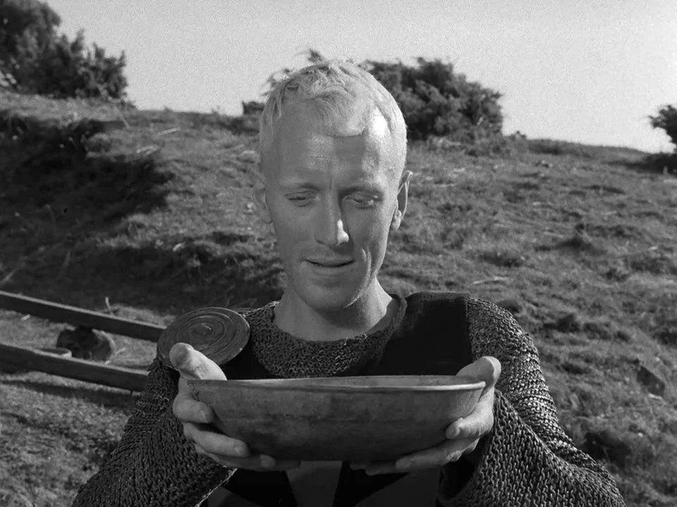The Seventh Seal (1957) movie still: medium — A grassy, uneven hill serves as the background under a bright overcast sky. A man in chainmail armor…