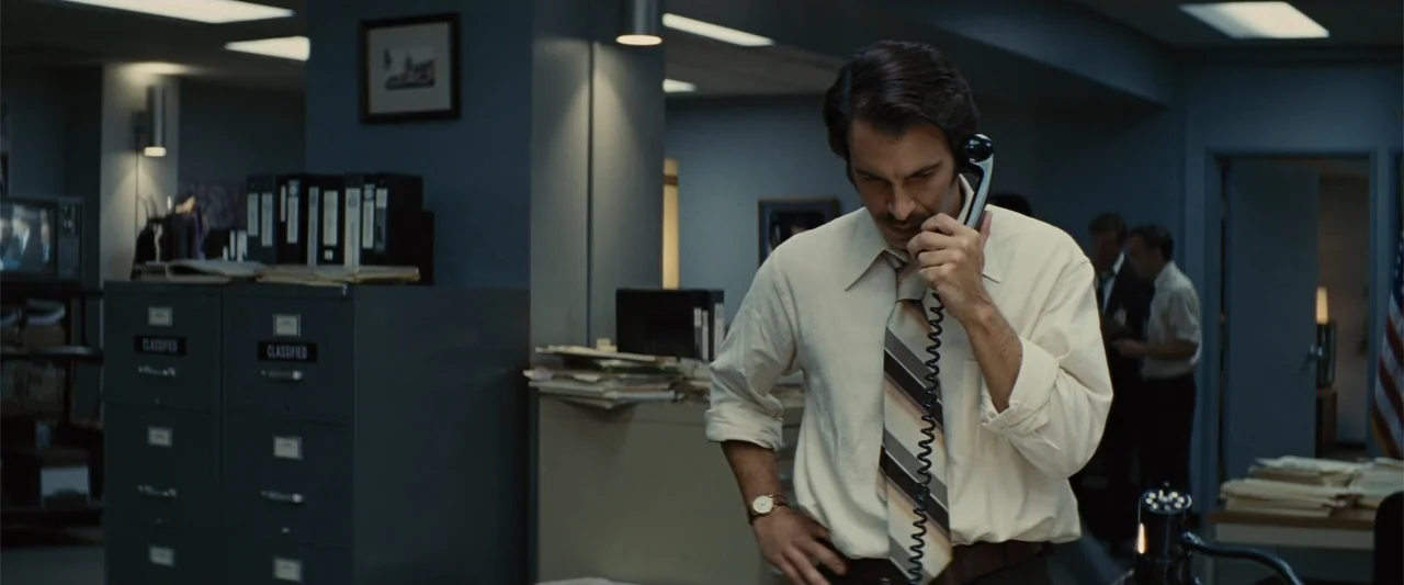 Argo (2012) movie still: medium — An office environment from the 1970s contains metal filing cabinets and stacks of documents. A man in a…