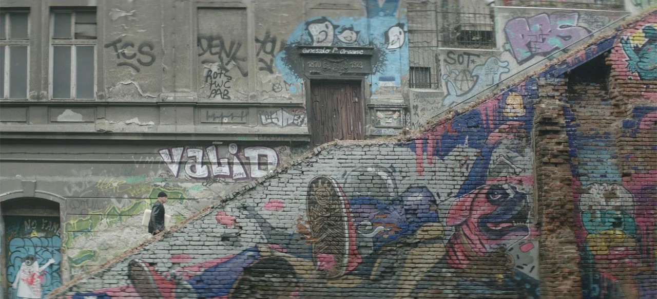 Morir un Poco (1967) movie still: wide — A weathered brick wall covered in extensive street art and graffiti lines an alleyway. A person in dark…