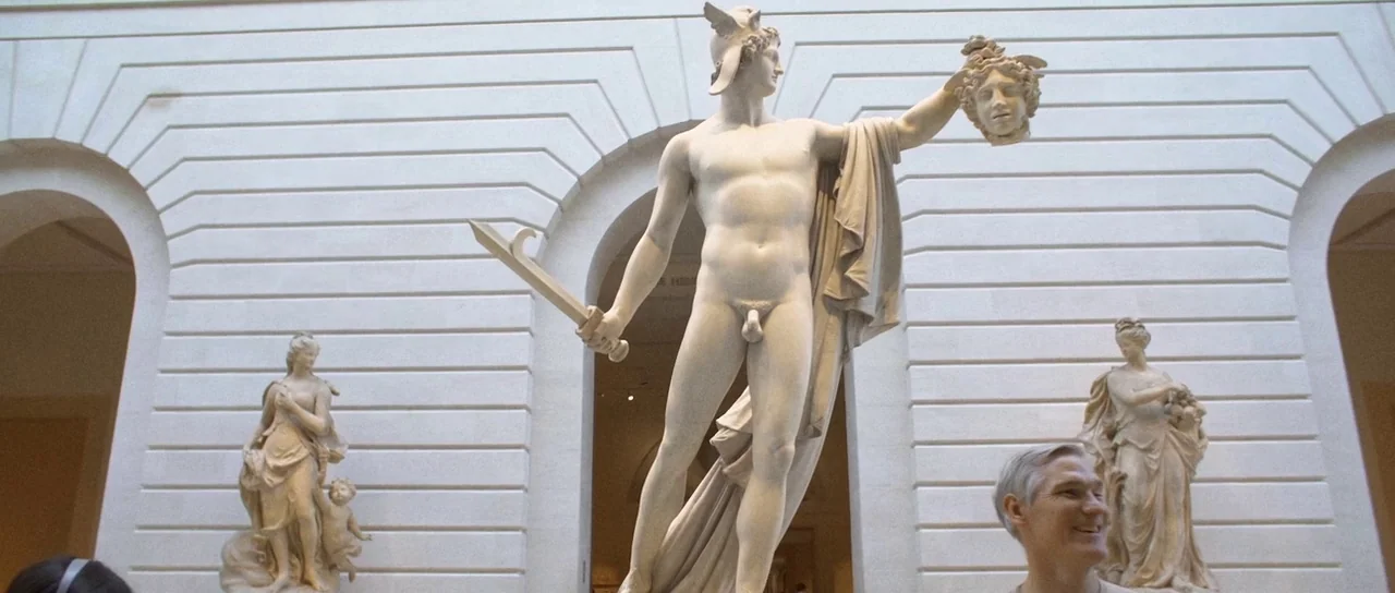 Depraved (2019) movie still: fullbody — A large marble statue of a male figure holding a head stands in a hall. White walls with arched…