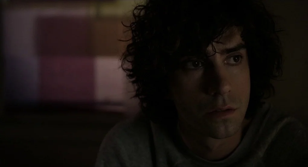 The Future (2011) movie still: closeup — A dimly lit room provides the backdrop for a close-up of a young man. The man sits motionless, looking…