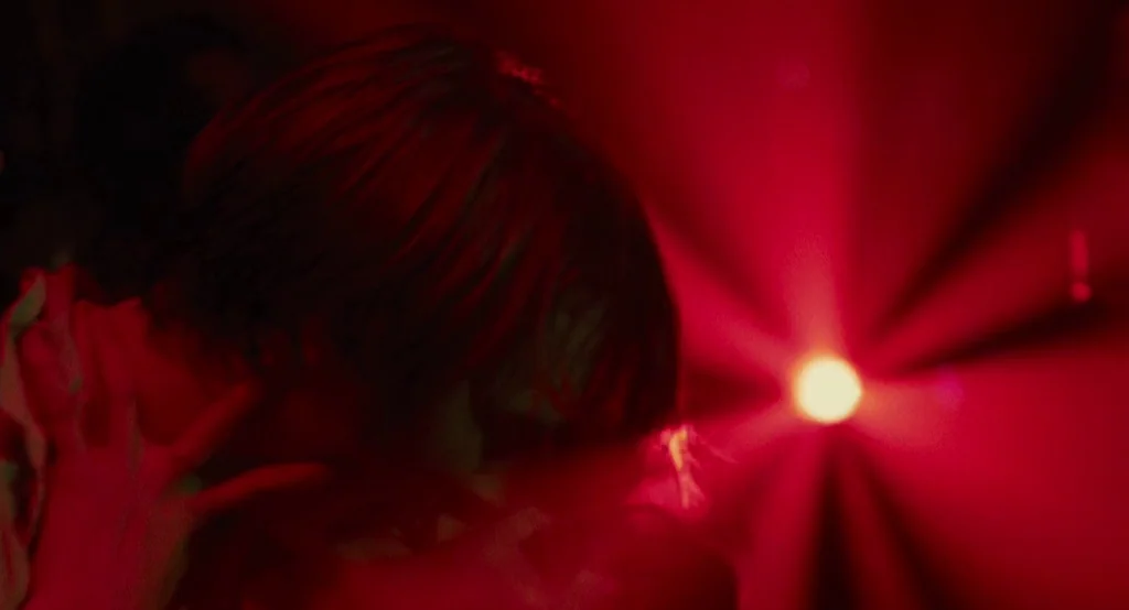 Ruby Sparks (2012) movie still: closeup — A dark space is illuminated by a single intense red light source and flares. A person with short hair is…