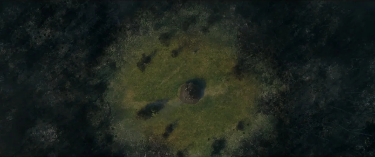 mother! (2017) movie still: establishing — A grassy clearing sits surrounded by a dense, dark forest canopy. An octagonal structure is centered in…
