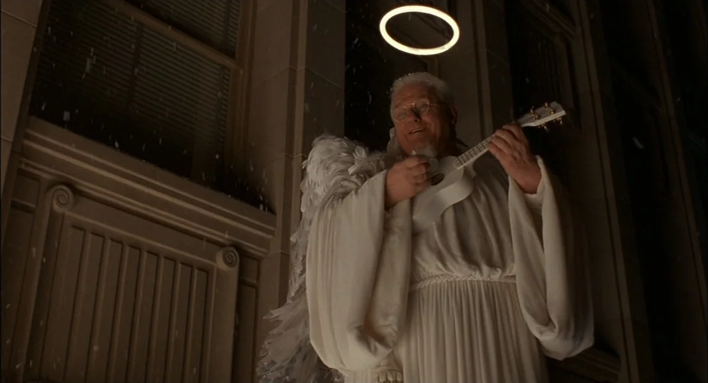 The Hudsucker Proxy (1994) movie still: medium — A man dressed as an angel stands in front of a dark building exterior. He wears white robes and…