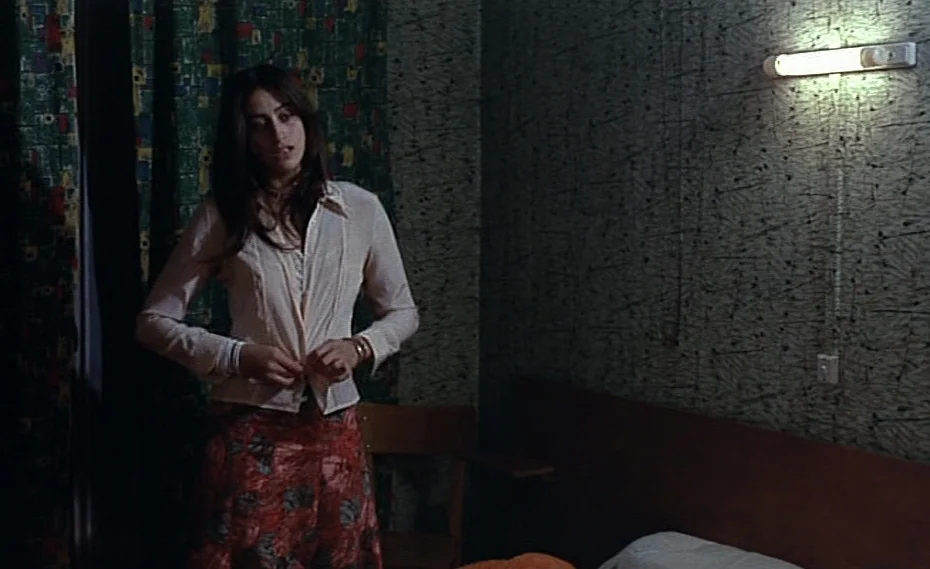 Bed & Board (1970) movie still: medium — A dim room features patterned wallpaper and a wall-mounted fluorescent light above a bed. A woman stands in…