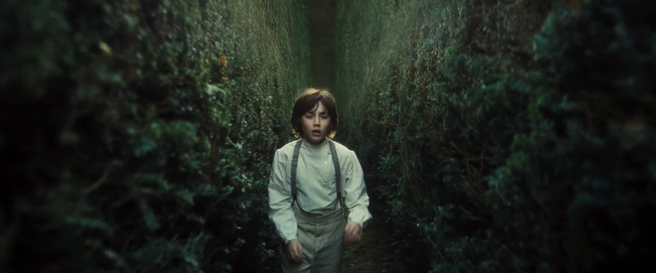 Anna Karenina (2012) movie still: medium — A tall, dense hedge maze features narrow dirt paths enclosed by thick, dark vegetation. A young boy in…