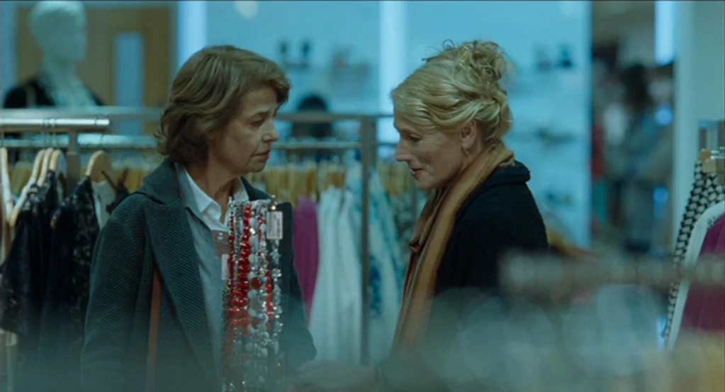 45 Years (2015) movie still: medium — A modern clothing store aisle is lined with racks of hanging garments on hangers. Two women stand in the aisle…