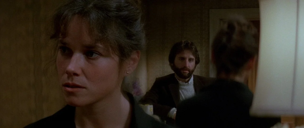 The Entity (1982) movie still: medium — A dimly lit room with patterned wallpaper contains a man sitting in the background. In the foreground, a…