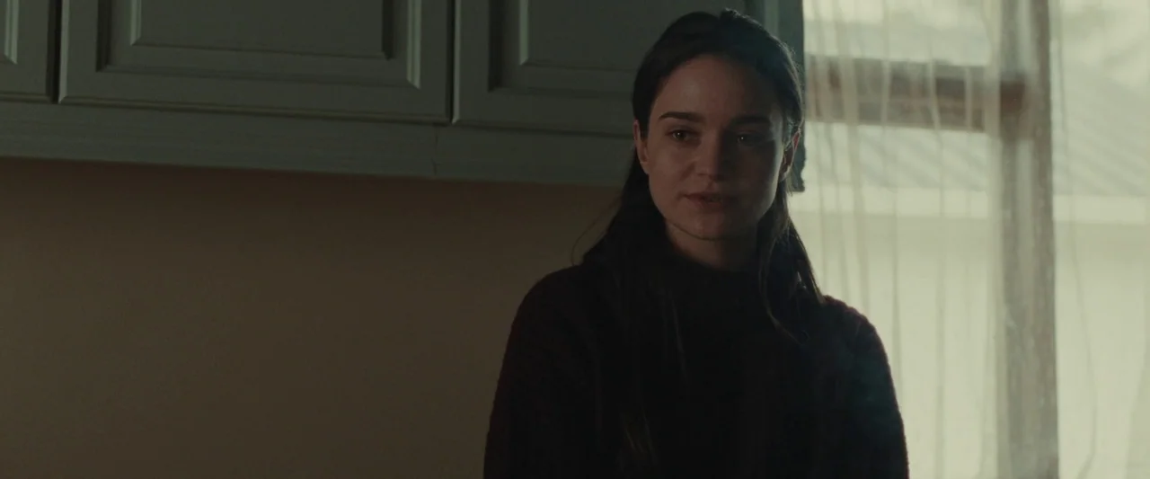 God’s Creatures (2022) movie still: medium — A residential room interior features light-colored cabinetry and a window with thin curtains. A woman…