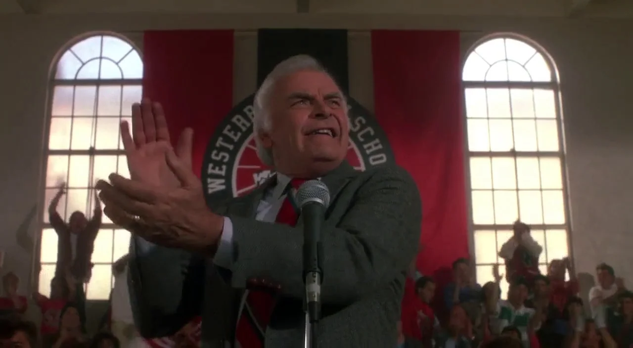 Heathers movie still: medium — A high school gymnasium interior features a large banner hanging on the back wall. An older man wearing a blazer…