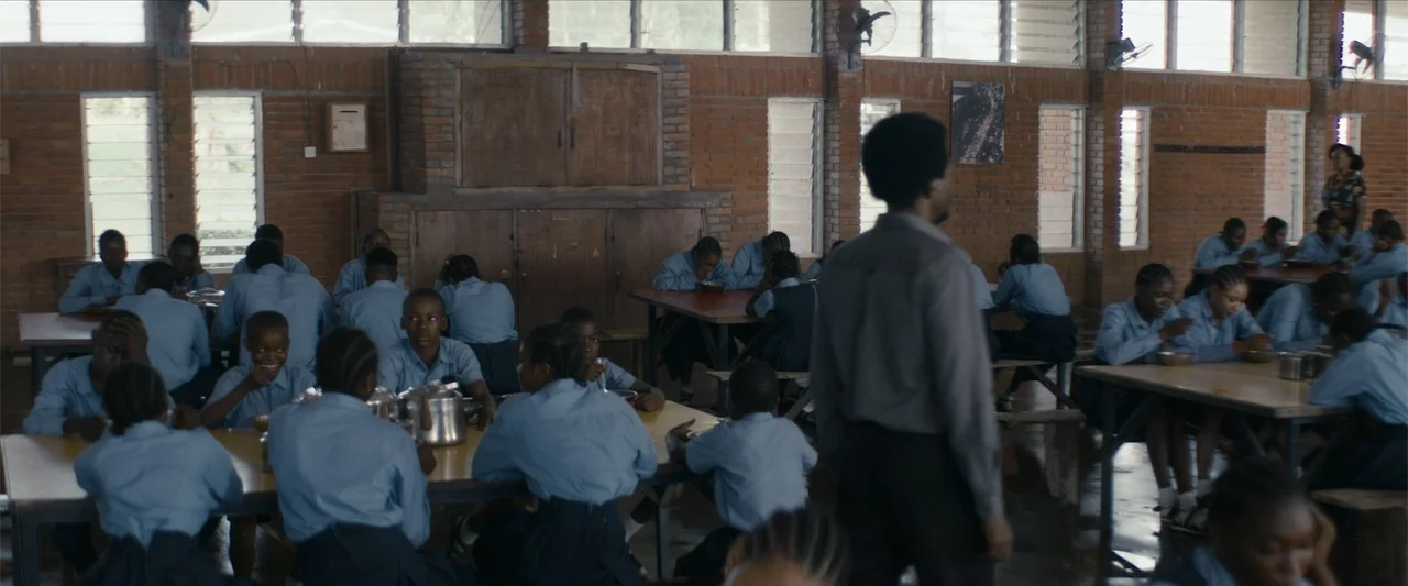 Ms. Kanyin (2025) movie still: wide — A large school dining hall features exposed red brick walls and wooden dining tables. A man stands in the…