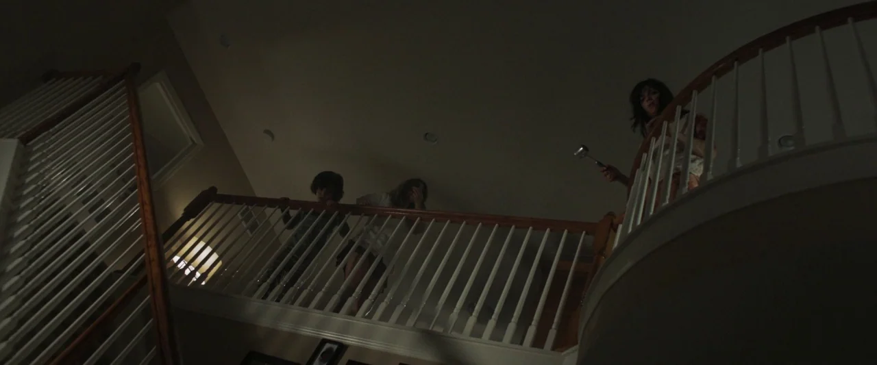 Mom and Dad (2017) movie still: wide — A residential staircase landing features white balusters and wooden railings set against dark walls. Three…