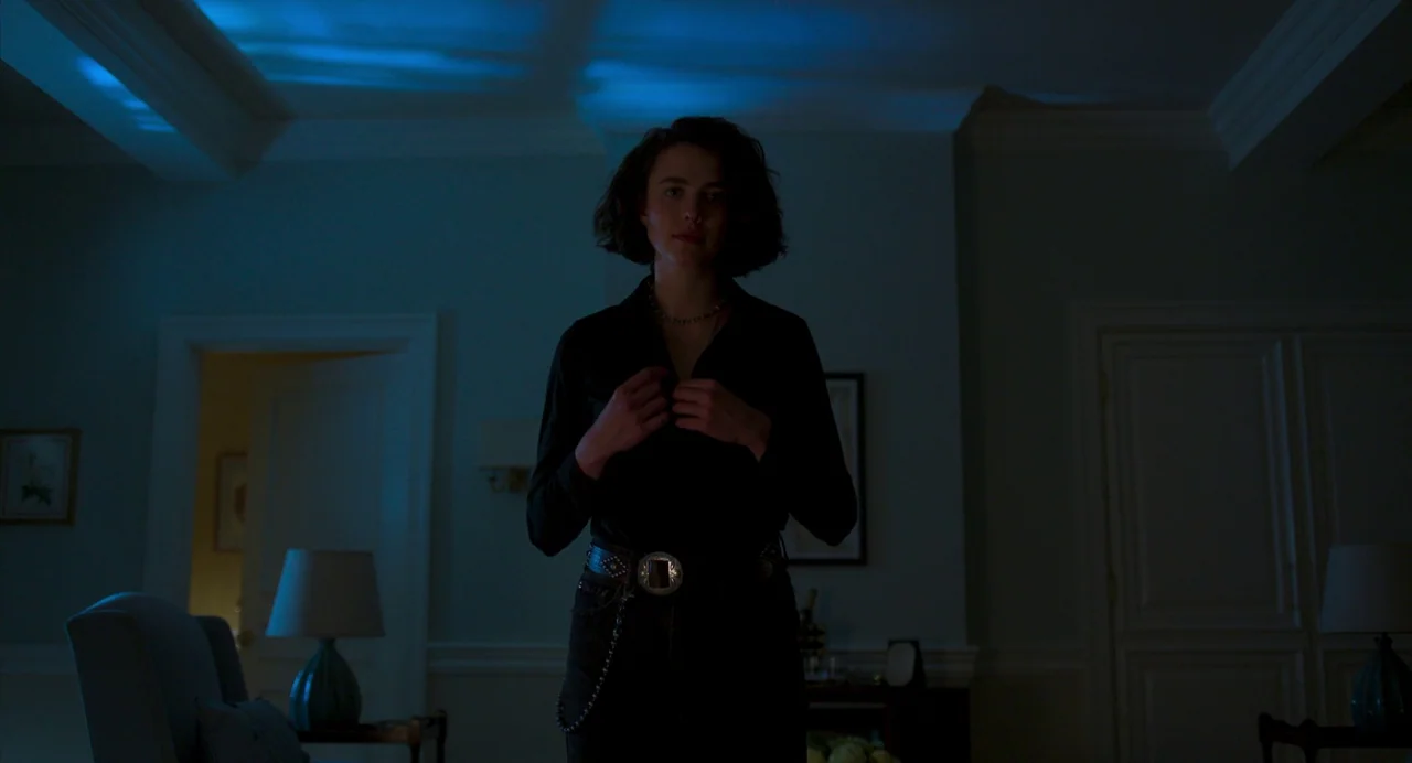 Drive-Away Dykes (2024) movie still: medium — A dimly lit room with blue ceiling reflections and closed white double doors. A young woman with dark…