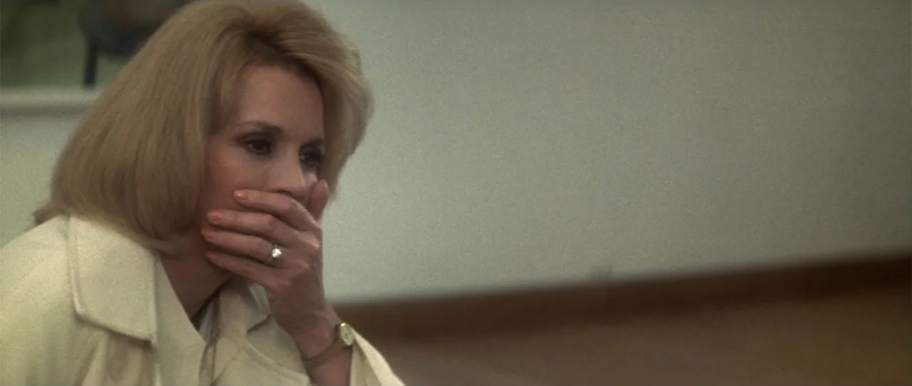 Dressed to Kill (1980) movie still: closeup — A woman in a cream coat sits in a sparse, brightly lit interior room. She holds her hand over her…