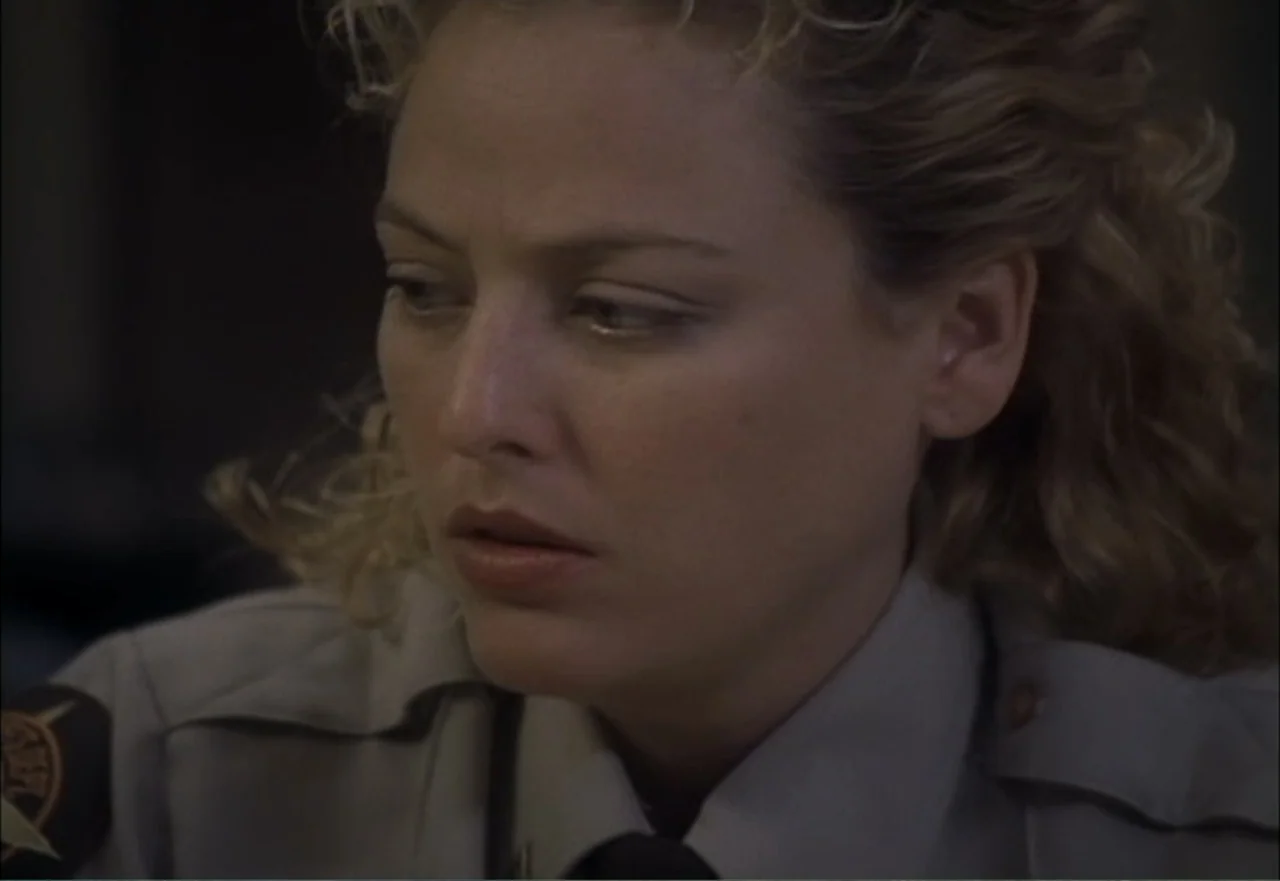 Ambushed (1998) movie still: closeup — A woman in a law enforcement uniform sits in a dimly lit office. She turns her head slightly while looking…