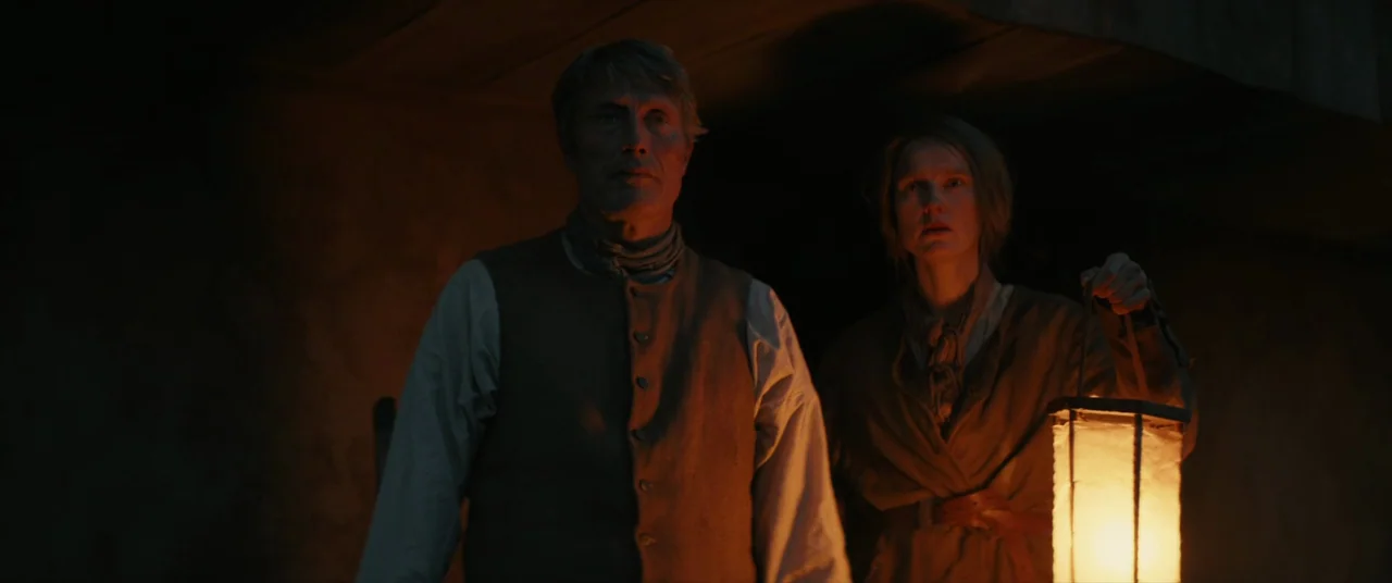 The Promised Land (2023) movie still: medium — A dark, cramped space with rough walls illuminated by a handheld candle lantern. A middle-aged man…