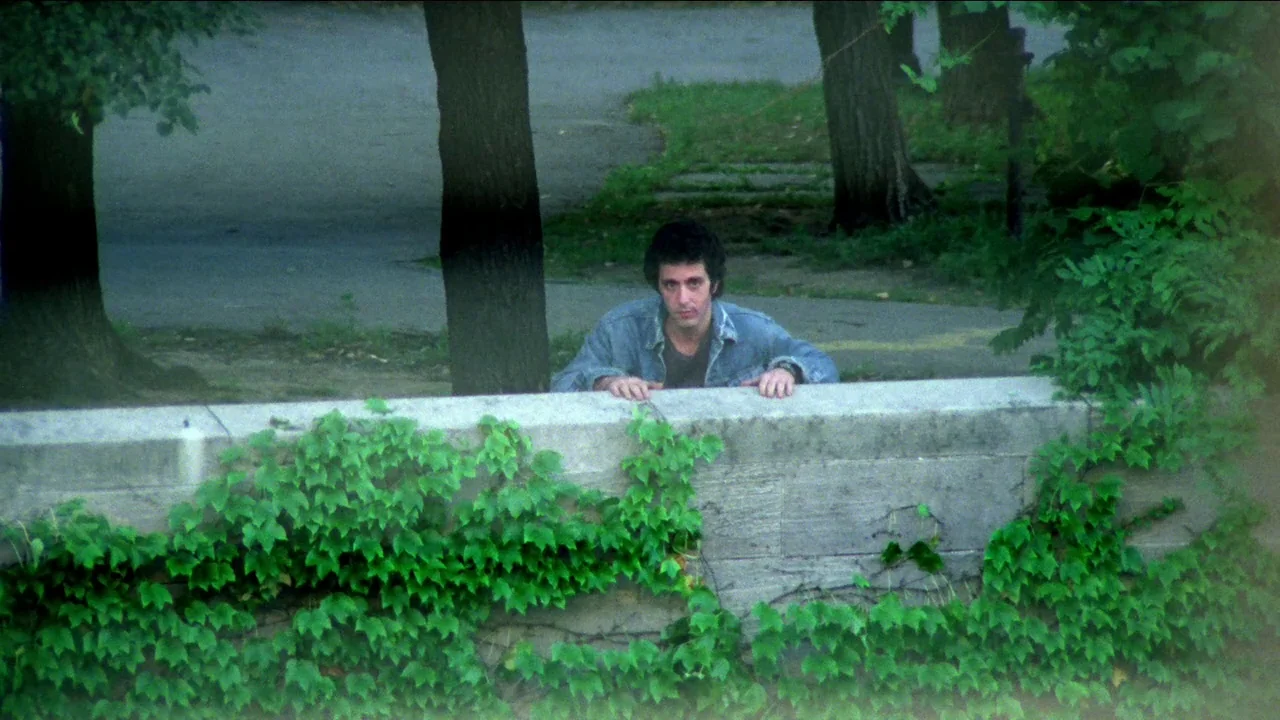 Cruising (1980) movie still: medium — A stone wall covered in climbing ivy sits in front of a tree-lined path. A man in a denim jacket rests his…