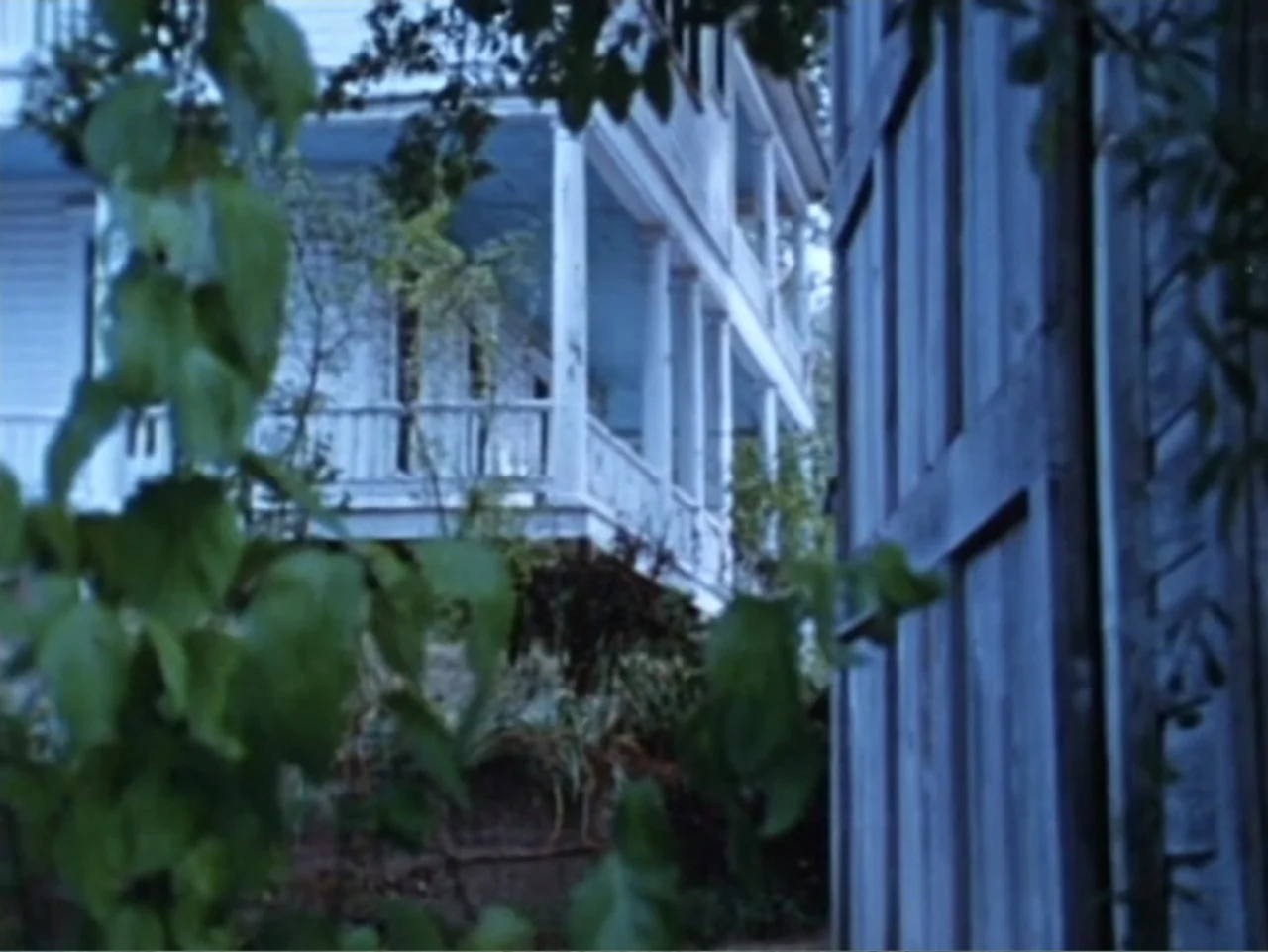 It's Alive (1974) movie still: wide — A white two-story wooden house with a porch is partially obscured by foliage. The structure sits in the dim…