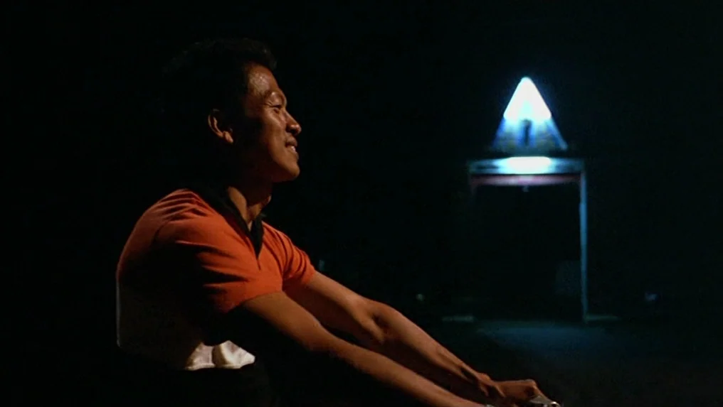 Tropical Malady (2004) movie still: medium — A dark night setting features a man riding a bicycle past a bright light. A glowing triangular sign…