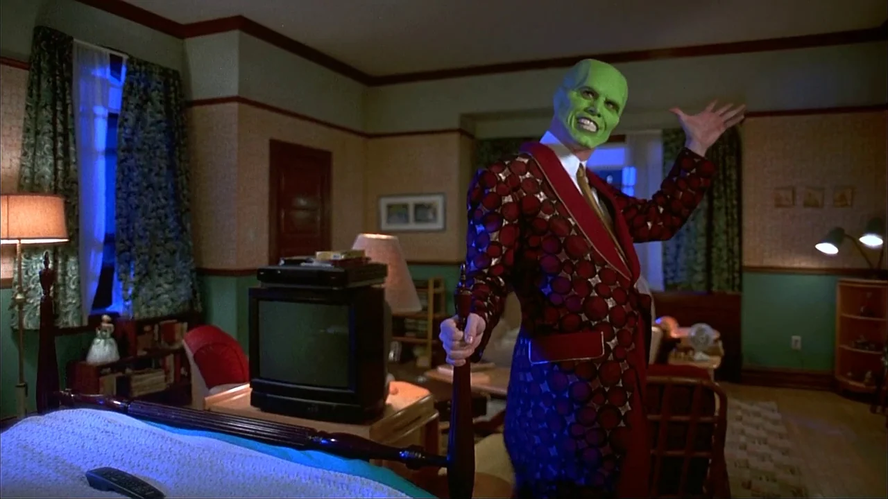 The Mask (1994) movie still: medium — A cluttered 1990s room features wallpaper and dark wooden furniture under dim indoor lights. A man with green…