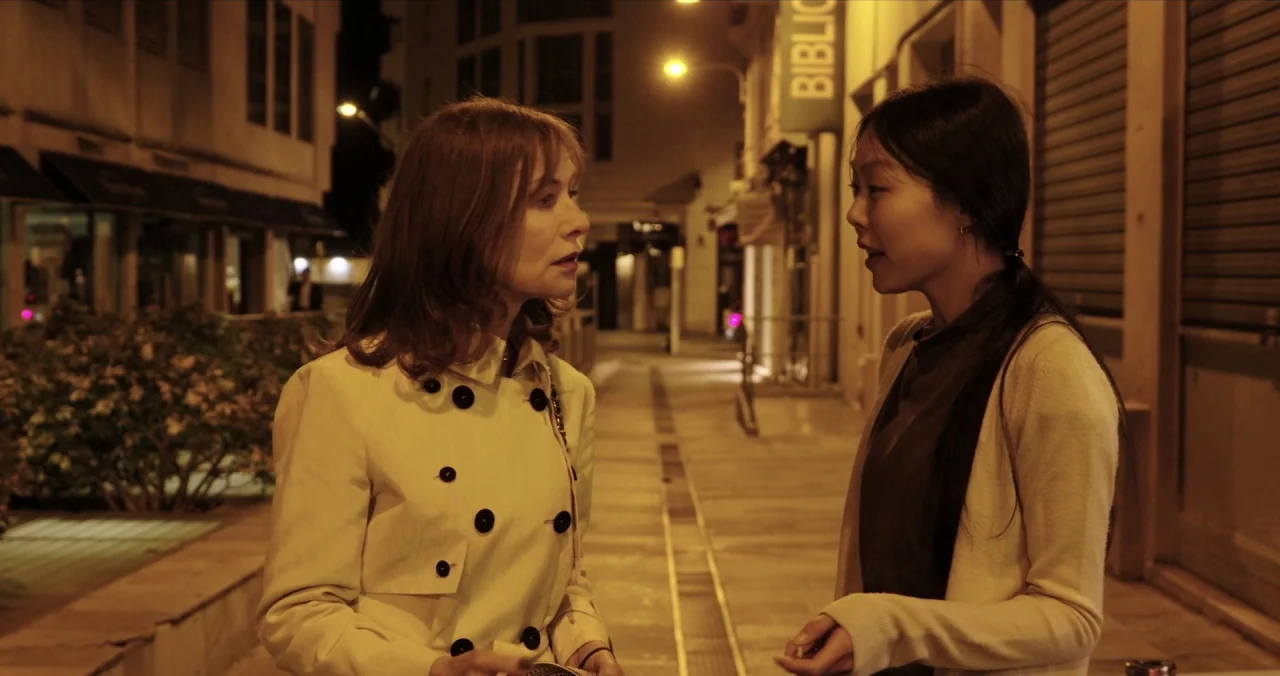 Claire’s Camera (2017) movie still: medium — A night time pedestrian area with tan pavement and modern urban buildings. Two women stand facing each…