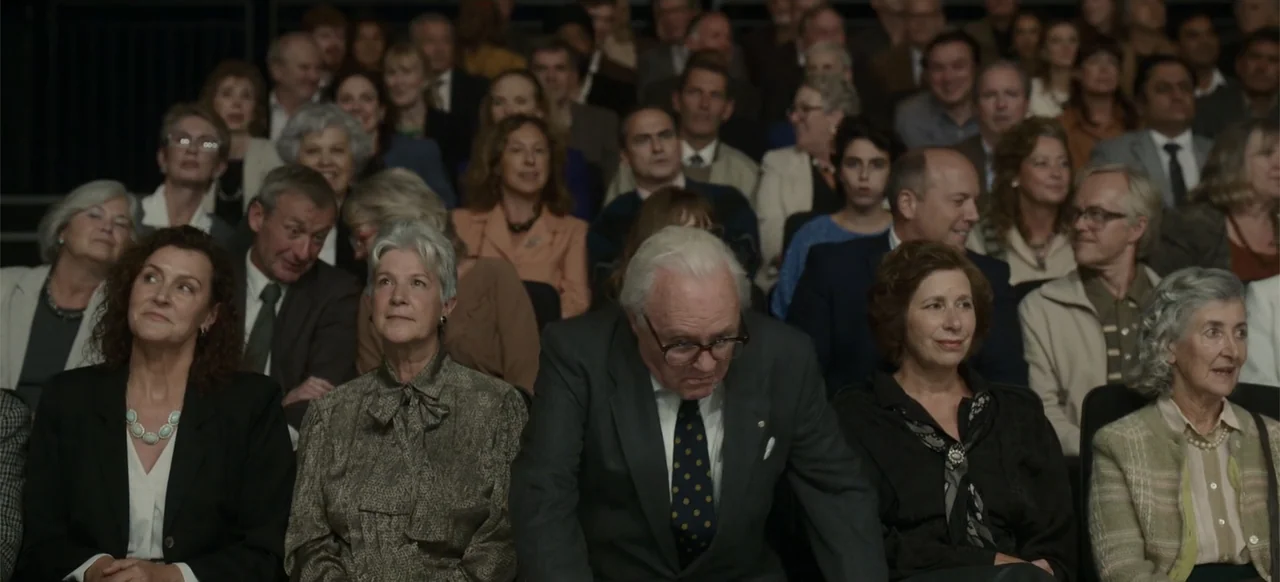 One Life (2023) movie still: medium — An indoor auditorium filled with a seated audience of older individuals in professional attire. The subjects…