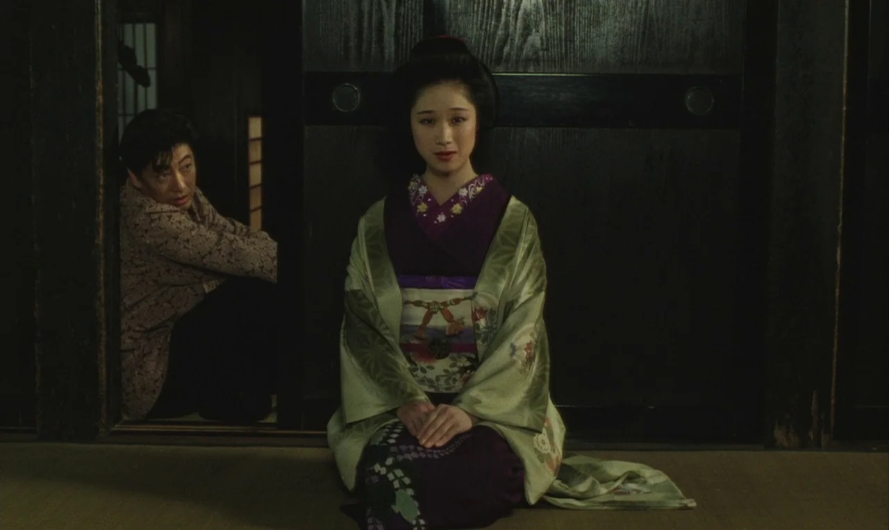 Yumeji (1991) movie still: medium — A traditional Japanese room features dark wooden panels and woven tatami mat flooring. A woman sits in the…