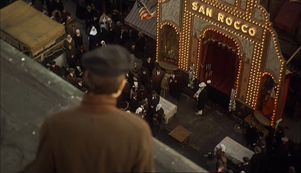 The Godfather Part II (1974) movie still: wide — A crowded urban street festival occurs at night under a large illuminated decorative arch. A man…