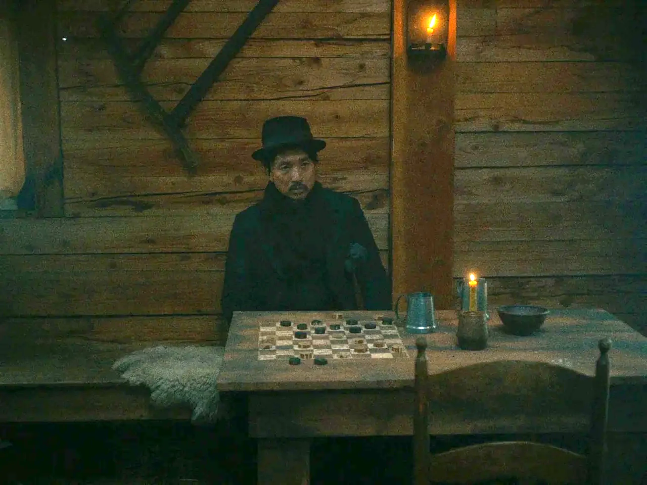 First Cow (2019) movie still: medium — A dimly lit log cabin features rough timber walls and a rustic wooden table. A man in a dark hat sits alone…