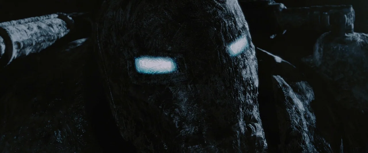Iron Man (2008) movie still: closeup — A dark, textured armored surface faces the camera directly in deep shadow. Two glowing horizontal rectangular…
