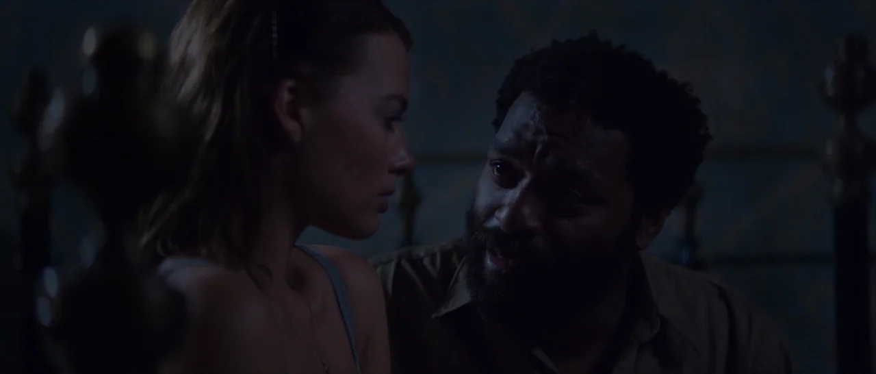 Z for Zachariah (2015) movie still: medium — A dimly lit room with dark blue walls features a pair of metal bed posts. A woman in a blue tank top…