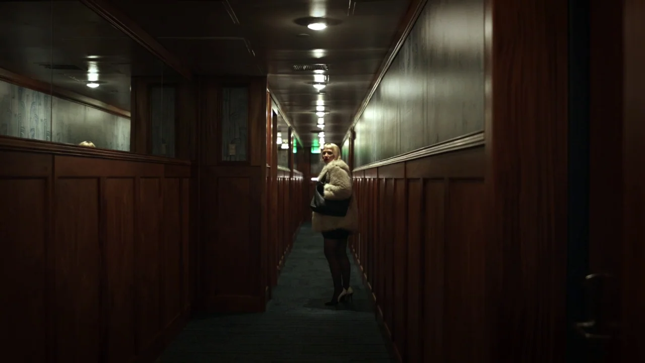 Piercing (2018) movie still: wide — A long corridor features dark wooden wainscoting and blue patterned wallpaper under recessed lighting. A woman…