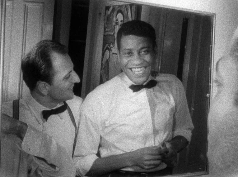 Shadows (1958) movie still: medium — A man stands behind another man while they look into a wall-mounted mirror. A framed piece of art hangs on the…