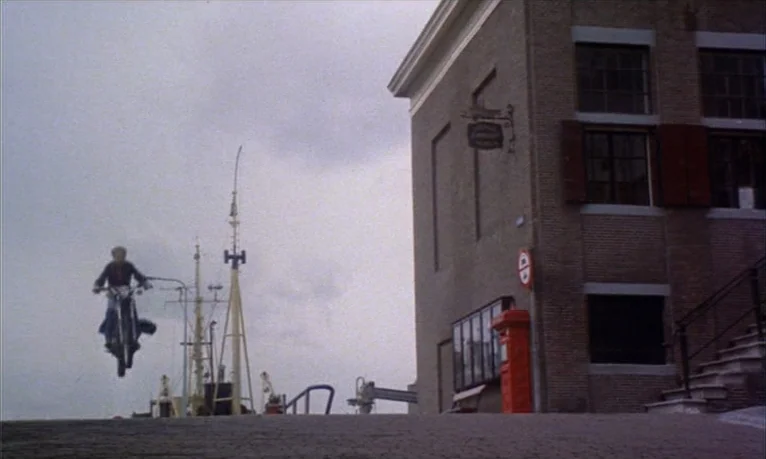 Spetters (1980) movie still: wide — A harbor area features a brick building and a mast of a ship. A motorcyclist executes a jump in the air against…