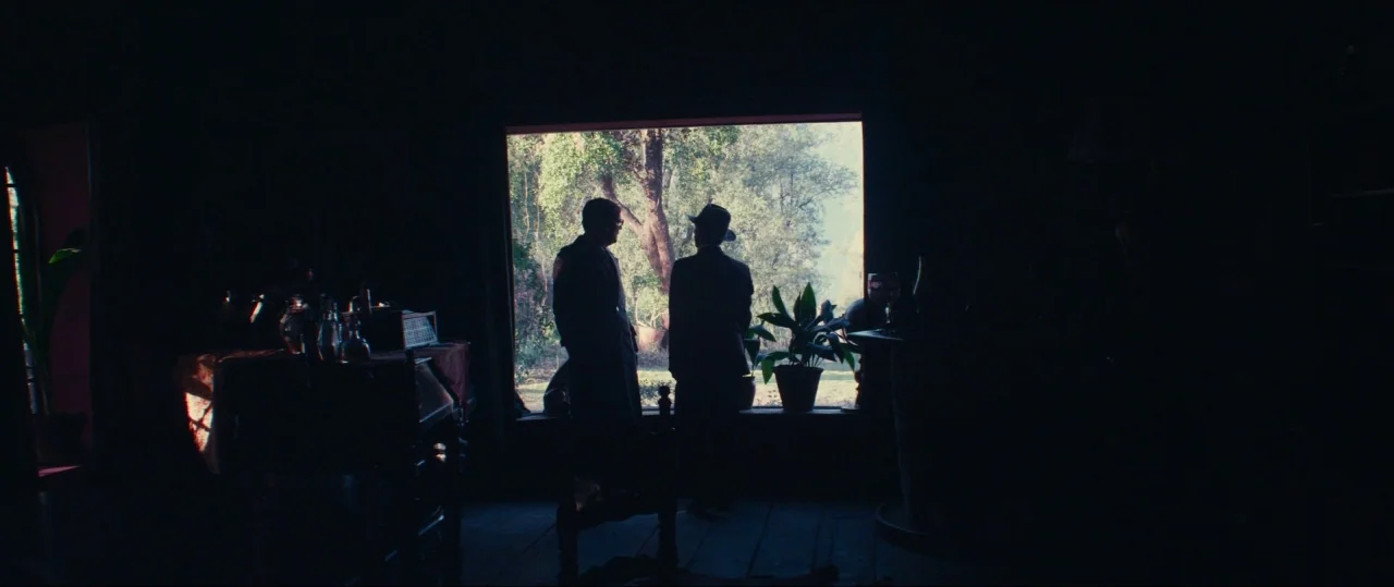 Neruda (2016) movie still: wide — A dark, wood-floored space contains a large window overlooking a lush, sunlit outdoor area. Two men in period…