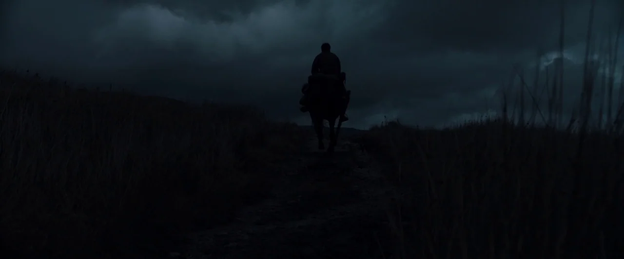 Black ’47 (2018) movie still: wide — A person on horseback moves slowly across a dark, grassy field under stormy clouds. The silhouette of the rider…