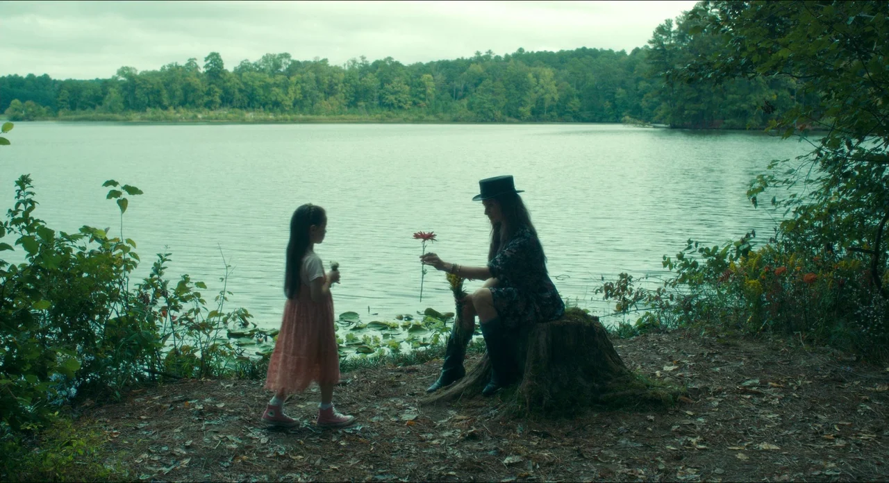 Doctor Sleep movie still: wide — A calm lake is surrounded by a dense forest under a cloudy overcast sky. A woman seated on a tree stump hands a…