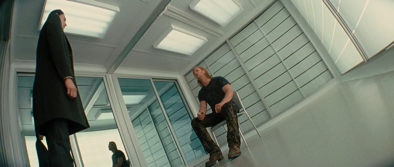 Thor (2011) movie still: wide — A clinical, windowless room features white modular panels and recessed fluorescent ceiling fixtures. Two men stand…